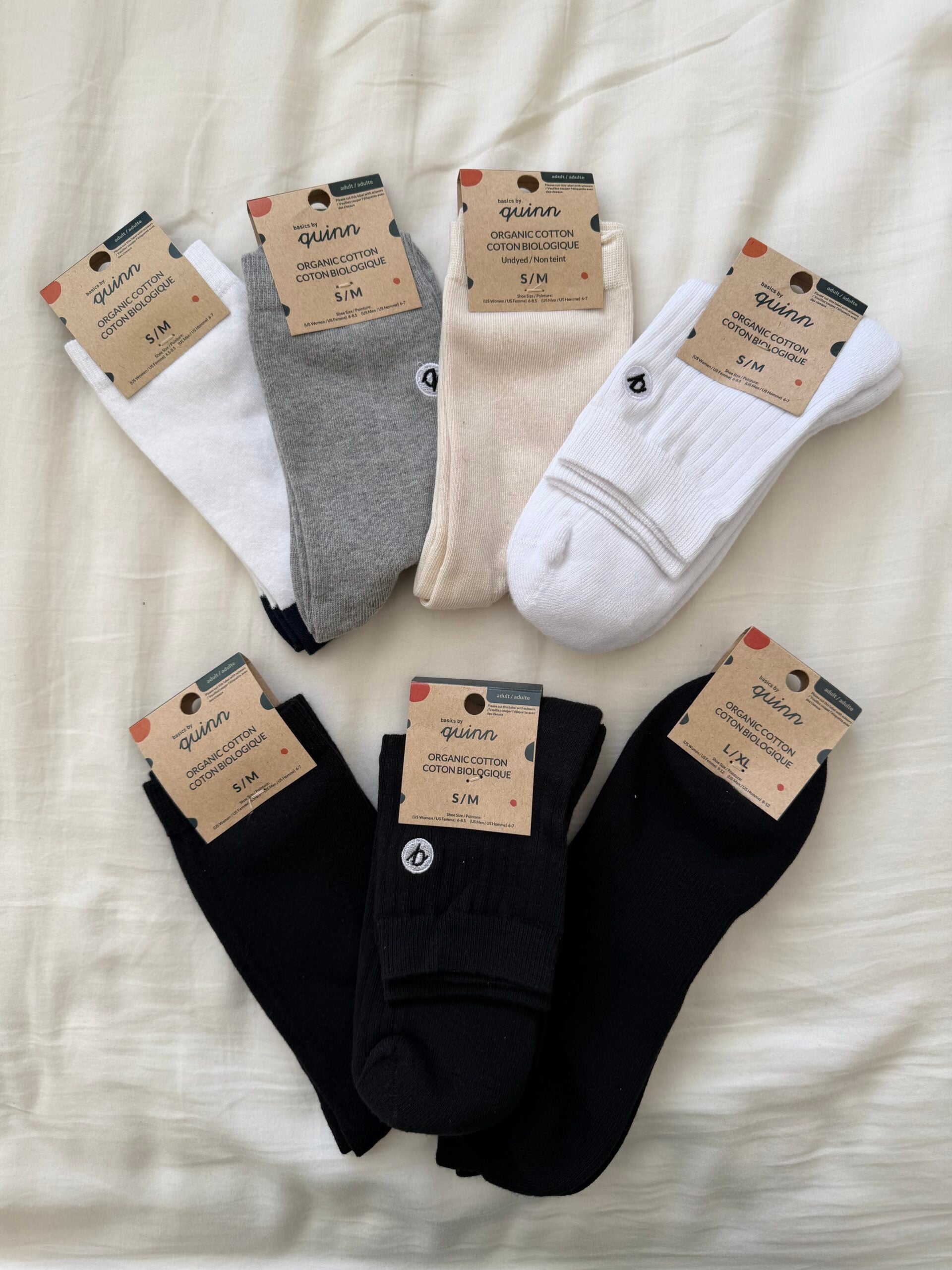 Six pairs of Quinn organic cotton socks in white, grey, cream, and black are arranged on a bed. Each pair is labeled with a brown cardboard tag indicating the size S/M.