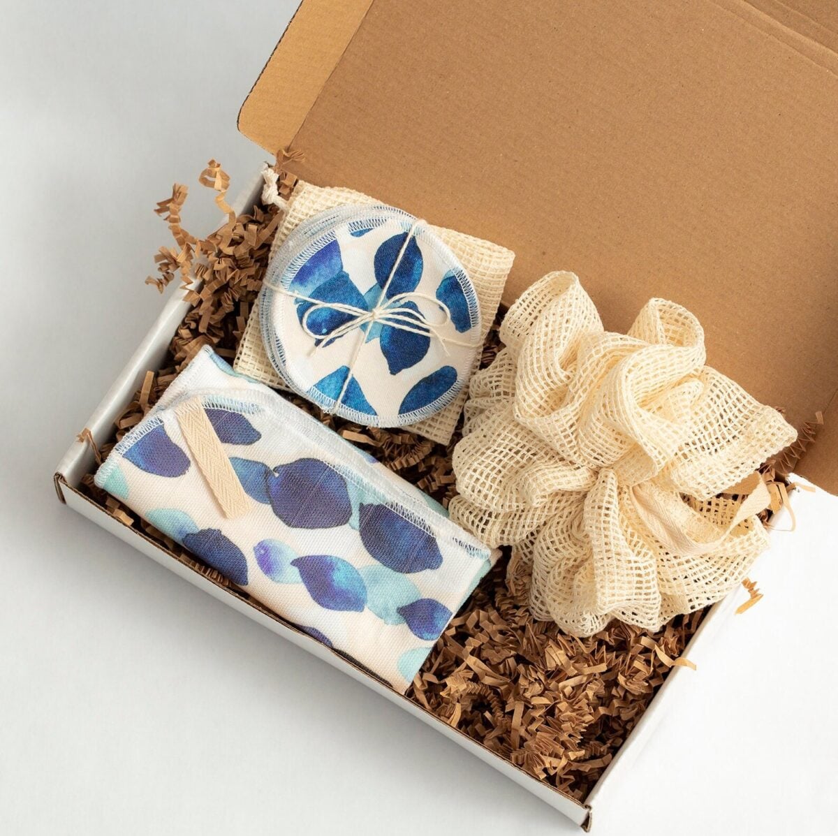 Eco-friendly gift box containing reusable makeup remover pads with blue leaf pattern and mesh produce bag nestled in kraft paper shred