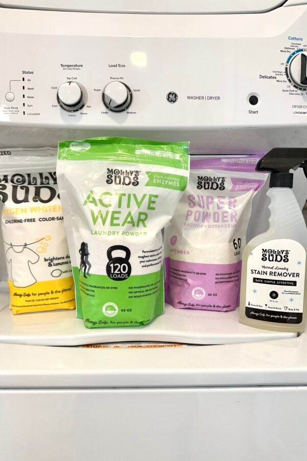 Four Molly’s Suds laundry products—Oxygen Whitener, Active Wear Laundry Powder, Super Powder Laundry Detergent, and Stain Remover—lined up on top of a white washer/dryer.