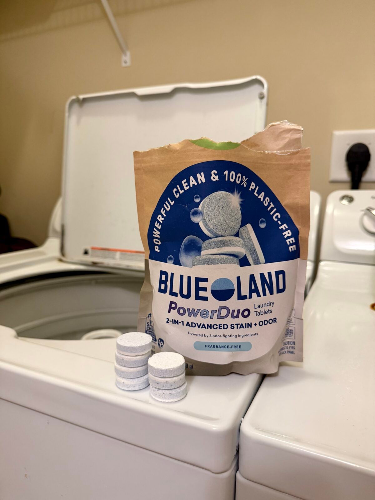 A bag labeled Blueland PowerDuo Laundry Tablets stands on a washing machine next to four stacked tablets. The bag highlights that the product is 2-in-1 and 100% plastic-free. The washing machine lid is open.