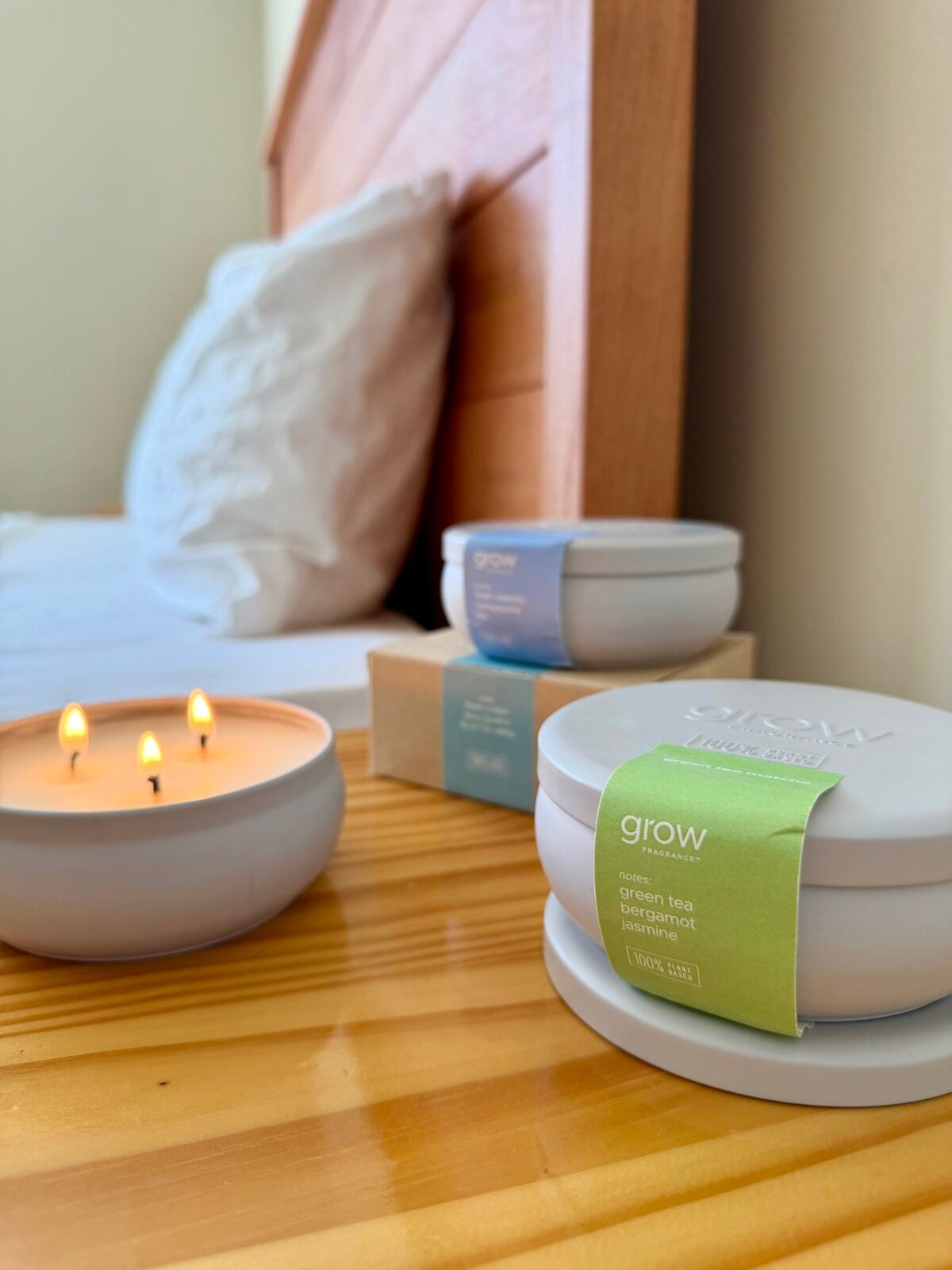 A lit three-wick candle sits on a wooden bedside table next to a bed with white pillows. Nearby are two closed round candle containers and a small cardboard box, all labeled grow with different scents.
