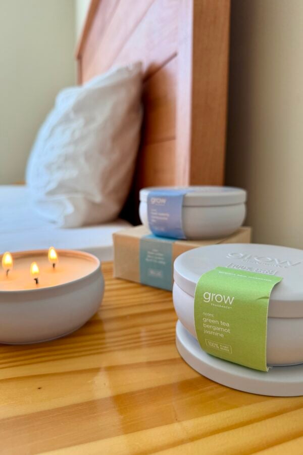 A lit three-wick candle sits on a wooden bedside table next to a bed with white pillows. Nearby are two closed round candle containers and a small cardboard box, all labeled grow with different scents.