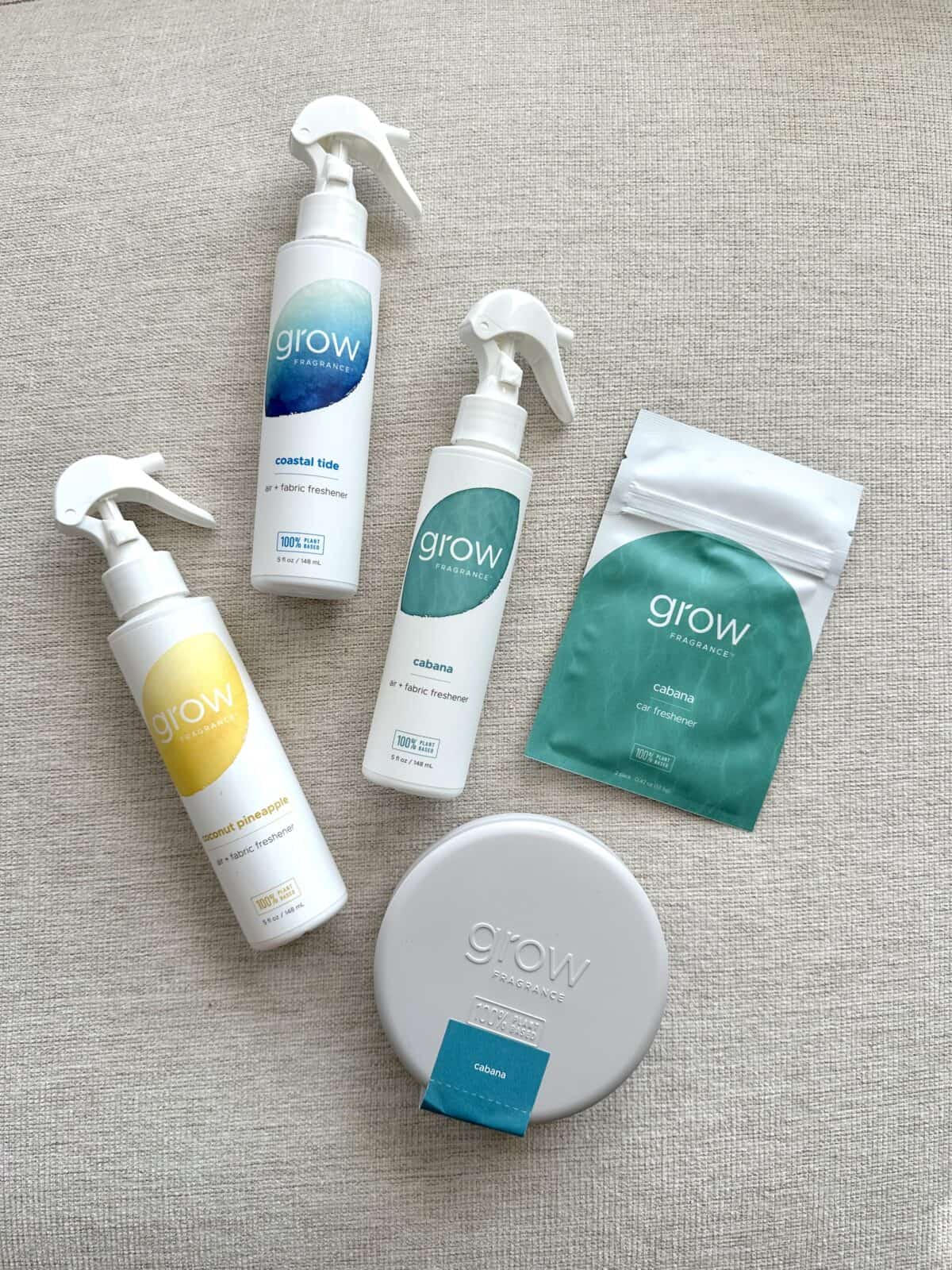 Flat lay of five Grow Fragrance products on a beige fabric surface, including three spray bottles (Coastal Tide, Cabana, and Coconut Pineapple), a Cabana car freshener packet, and a Cabana scented tin.