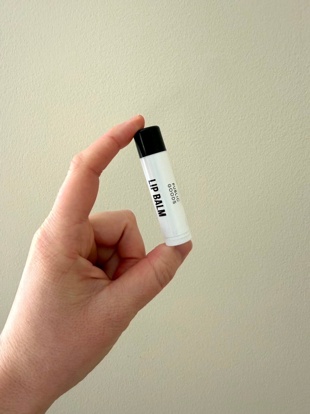 Hand holding Public Goods lip balm tube with black cap against beige wall