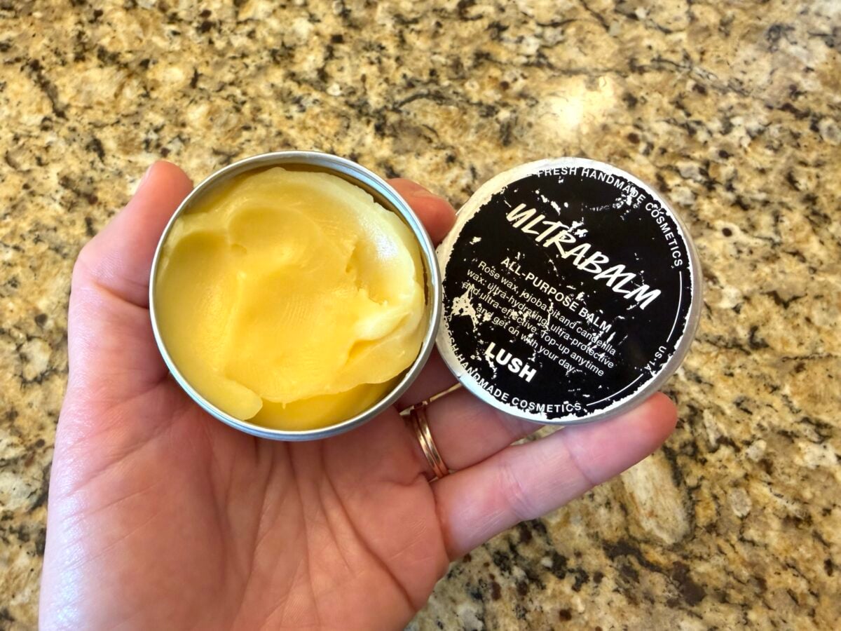 A hand holds an open tin of Lush Ultrabalm all-purpose balm, showing the yellow balm inside. The black and white lid rests on the hand, and the background is a granite countertop.