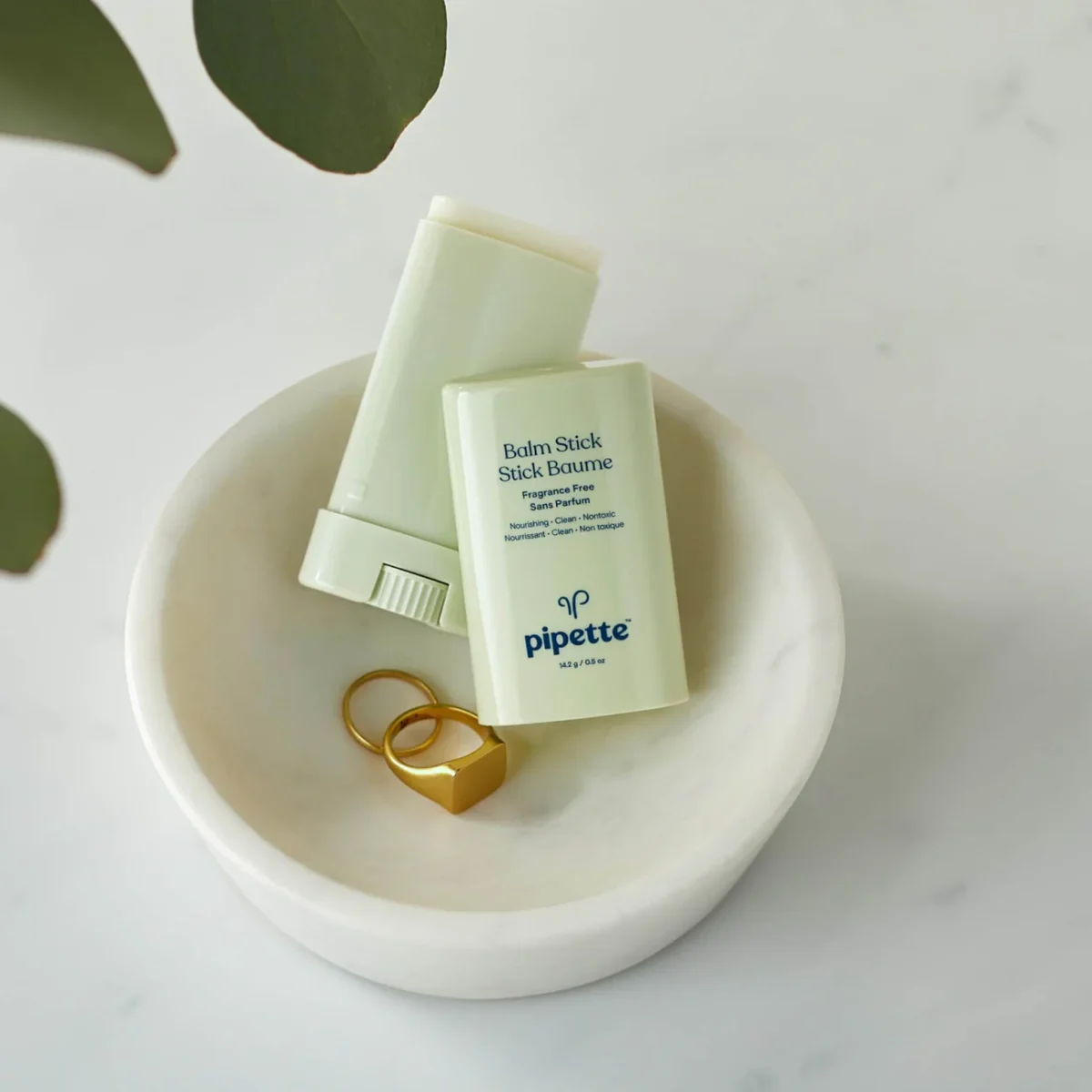 A white marble dish holds two light green Pipette Balm Stick containers and two gold rings, one square-shaped, on a white surface with green leaves partially visible at the top left.