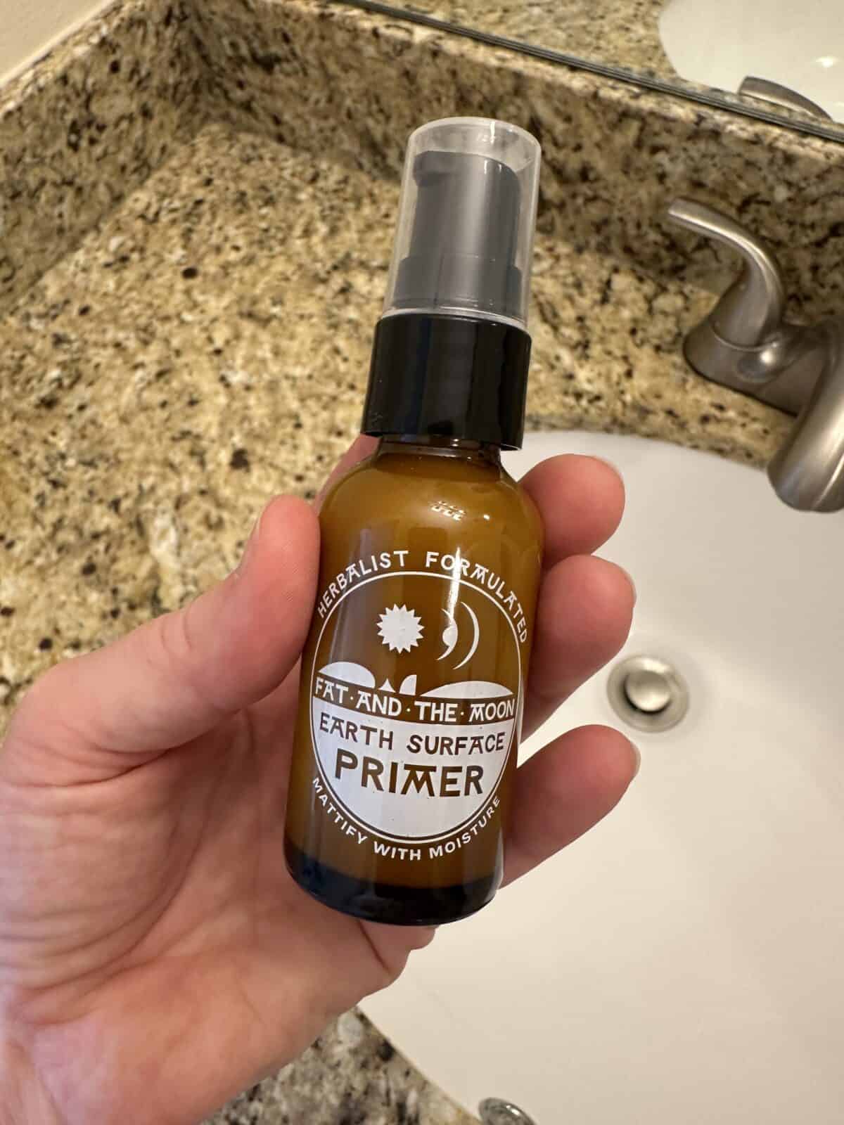 
A person is holding a small, brown glass bottle of Fat and the Moon Earth Surface Primer over a white bathroom sink with a marble countertop.
