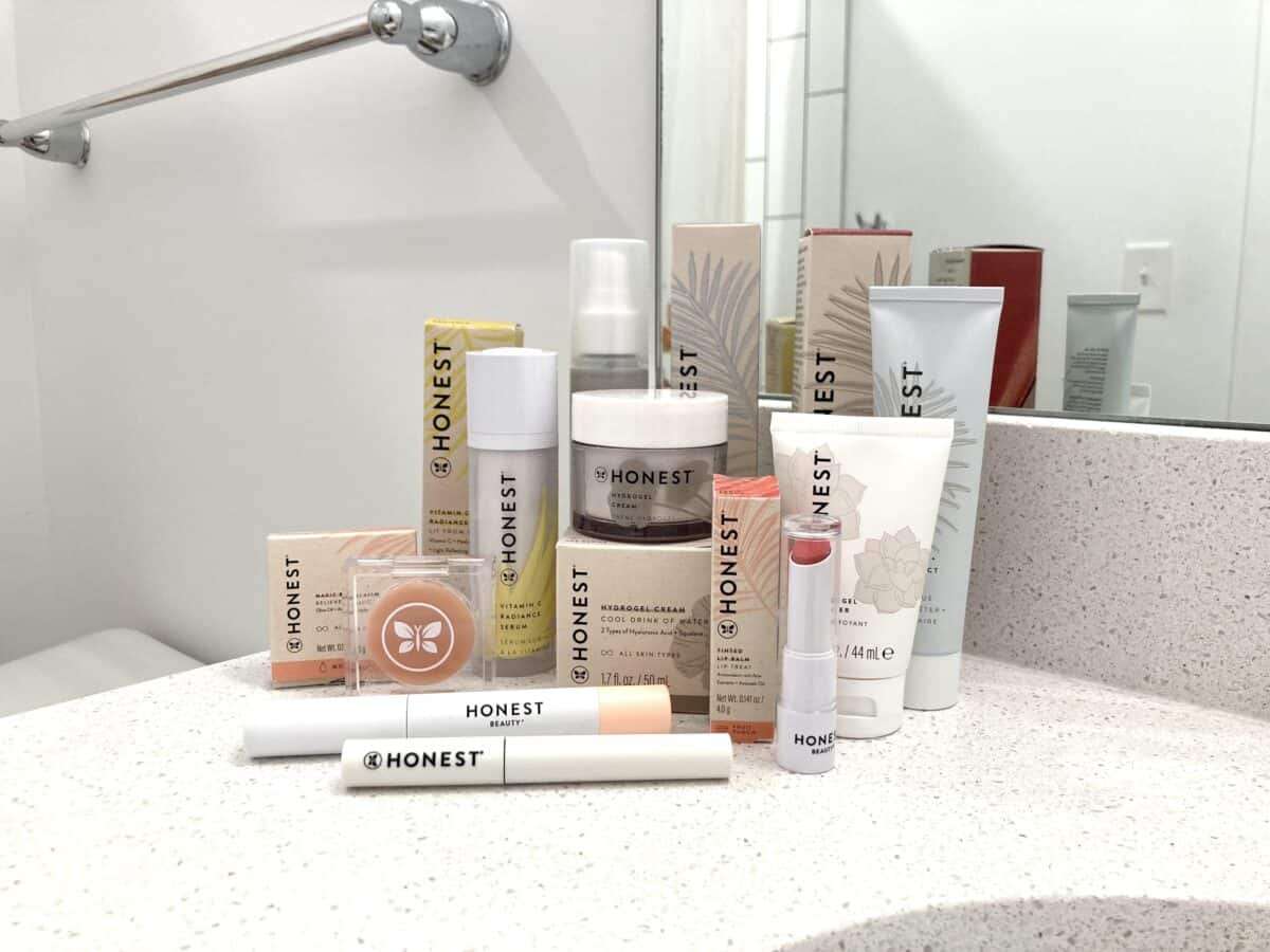 A collection of Honest Beauty skincare and makeup products, including serums, creams, lip color, and mascara, is arranged on a white bathroom counter in front of a mirror.