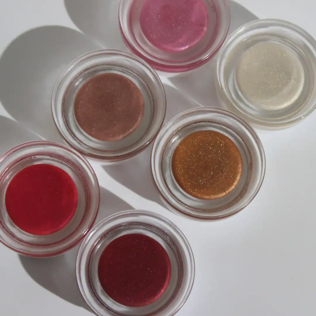 An overhead shot displays six small glass pots containing shimmering natural play makeup from Honey in shades of pink, red, bronze, gold, and clear gloss on a white surface.