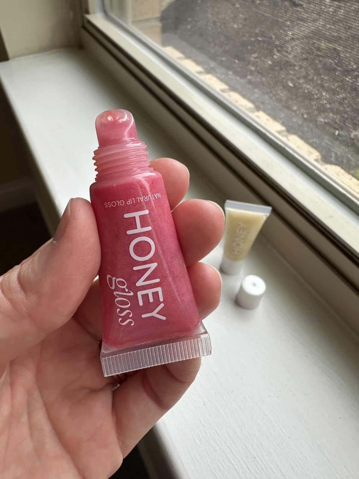 A person's hand holds a pink squeeze tube of Honey Gloss lip gloss near a sunlit window sill, with a second tube of lip gloss visible and out of focus in the background.