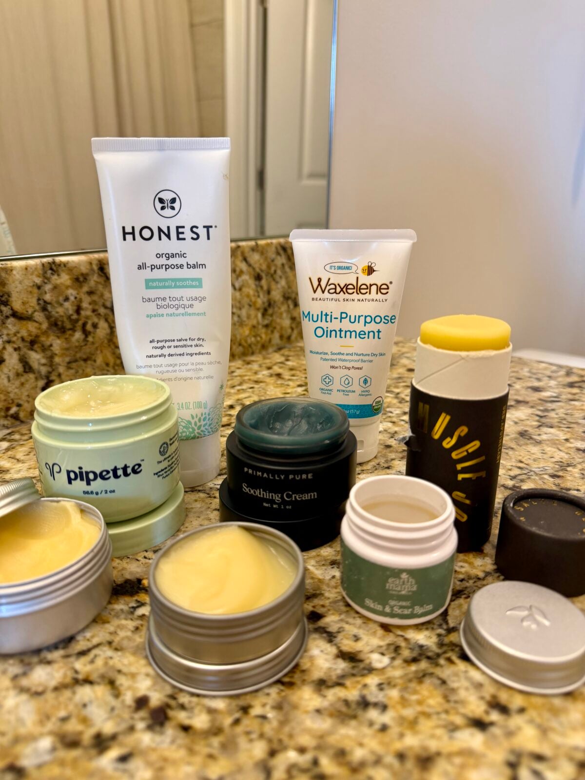 A selection of skincare products, including balms, ointments, and creams, arranged on a granite bathroom counter near a mirror. Brands such as Honest, Waxelene, and Pipette are visible.