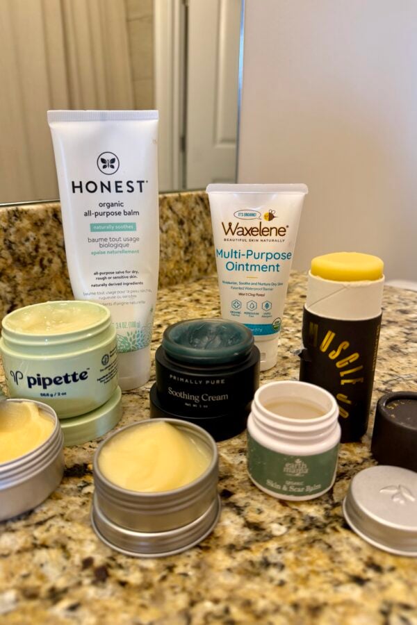 A selection of skincare products, including balms, ointments, and creams, arranged on a granite bathroom counter near a mirror. Brands such as Honest, Waxelene, and Pipette are visible.