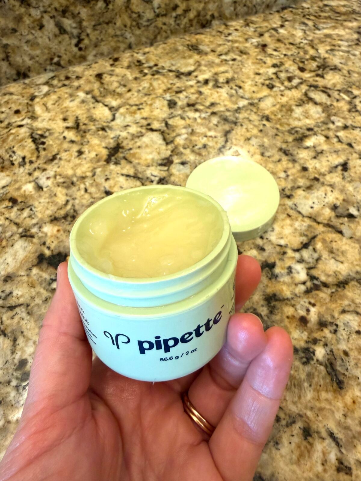 A hand holds an open jar of Pipette balm on a speckled granite countertop, with the lid resting nearby. The balm inside the jar is smooth and pale yellow.