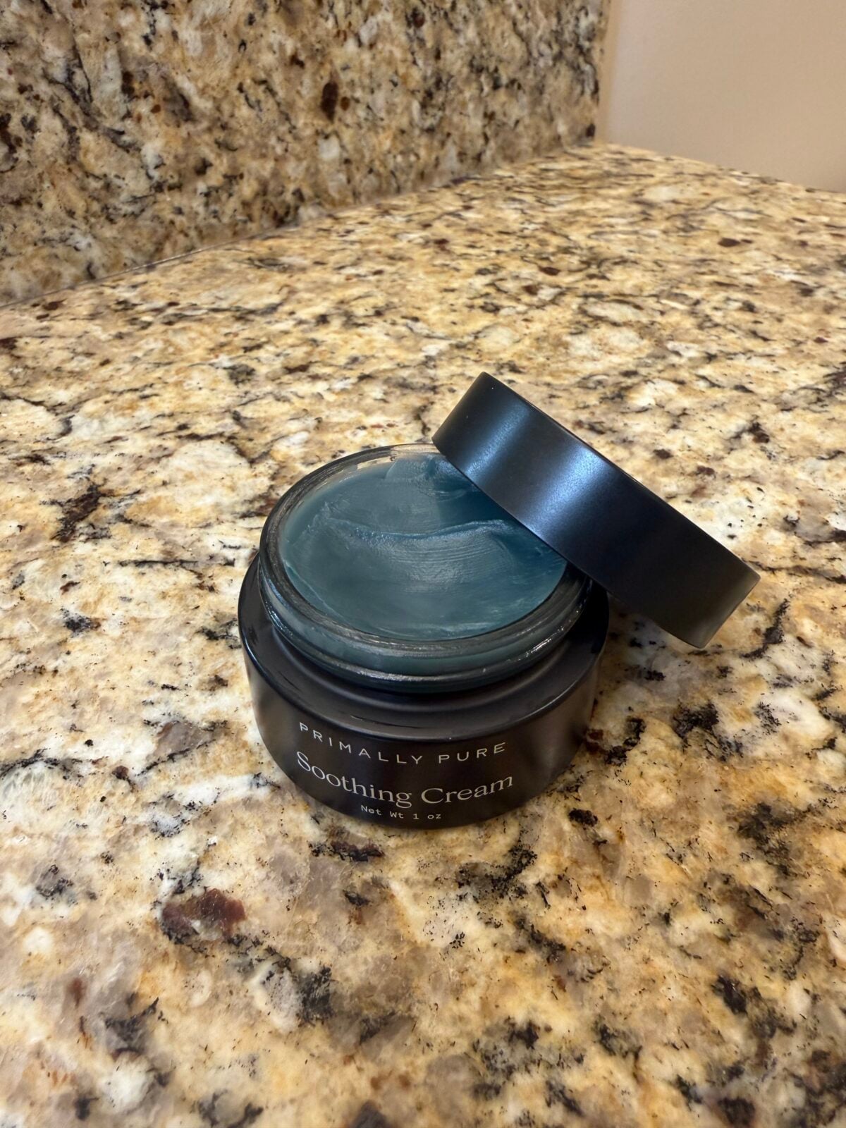 An open black jar labeled Primally Pure Soothing Cream sits on a speckled granite countertop. The lid is resting askew on top of the jar, showing the blue cream inside.