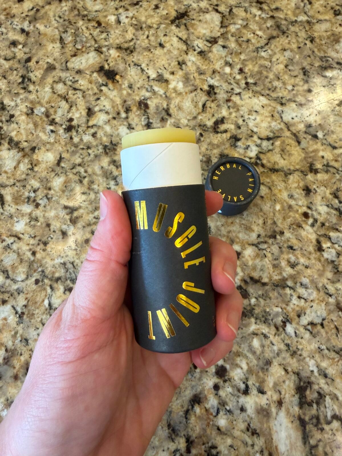 A hand holds a cylindrical container labeled MUSCLE JOINT with a yellow balm exposed at the top. A round container lid with HERBAL SALVE text lies on a speckled granite countertop in the background.