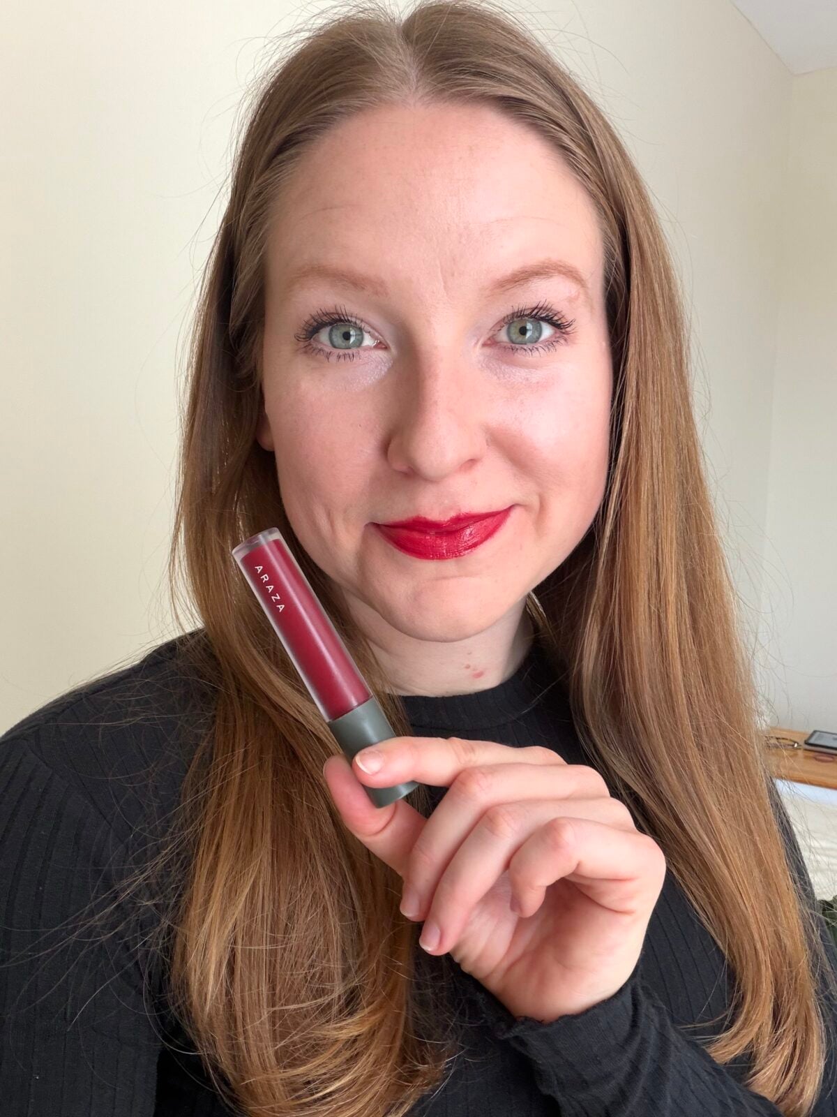 The photo shows a smiling woman with long brown hair wearing red lipstick and holding a tube of Araza brand lip color.