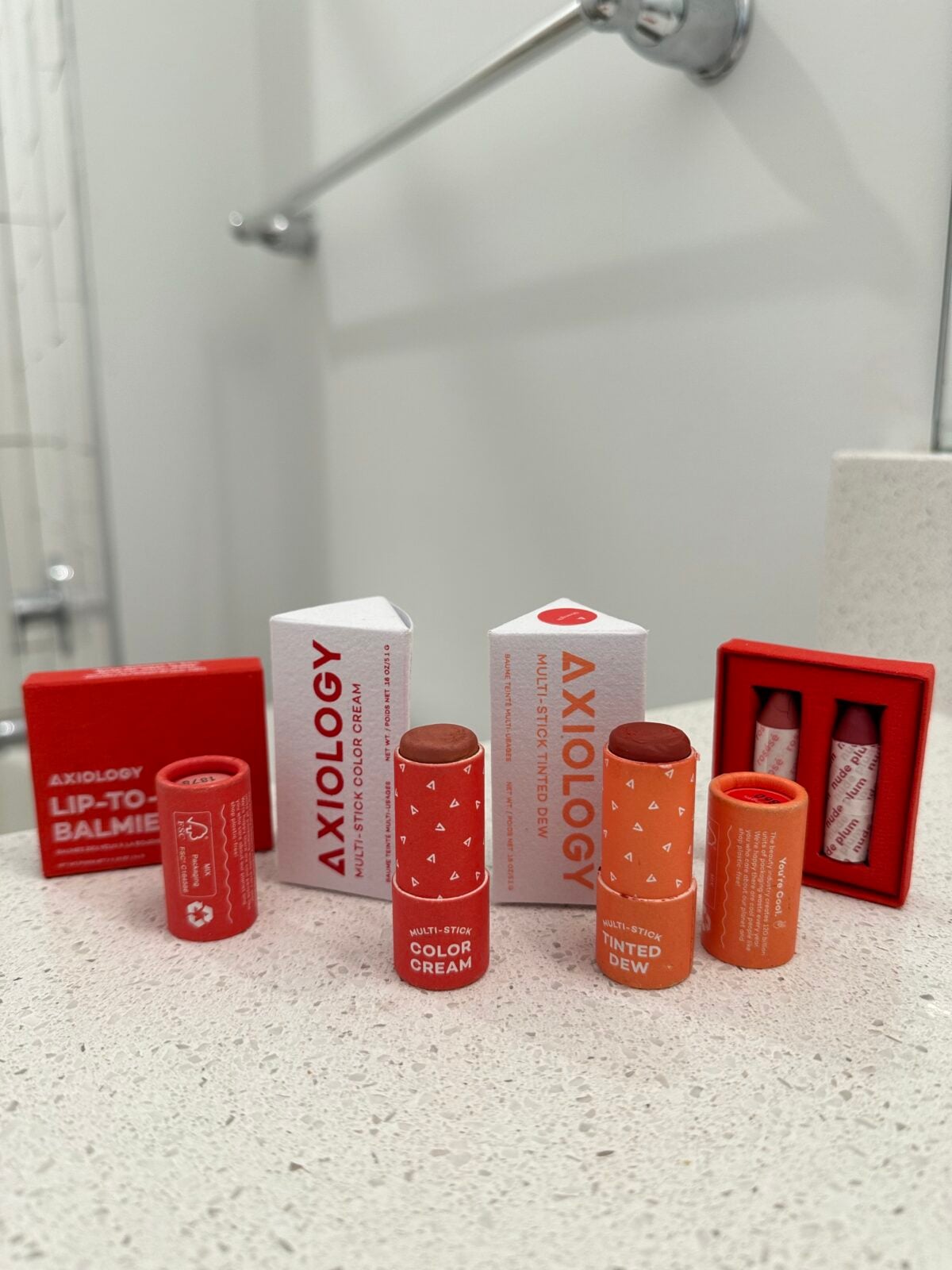 The photo shows an assortment of Axiology beauty products, including "Multi-Stick Color Cream," "Multi-Stick Tinted Dew," and "Lip-to-Balmie" tubes and boxes, arranged on a speckled white countertop.