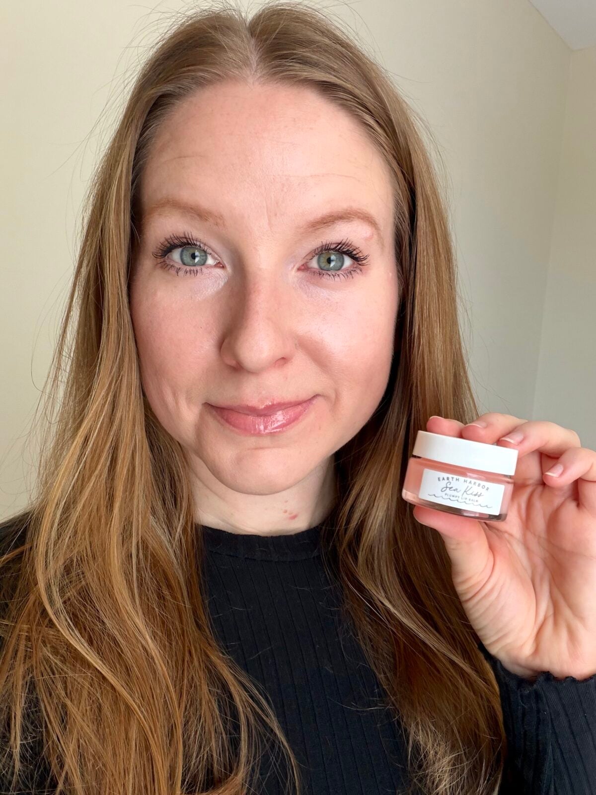 The photo shows a smiling woman with long brown hair holding a small glass jar of Earth Harbor's plumpy lip balm