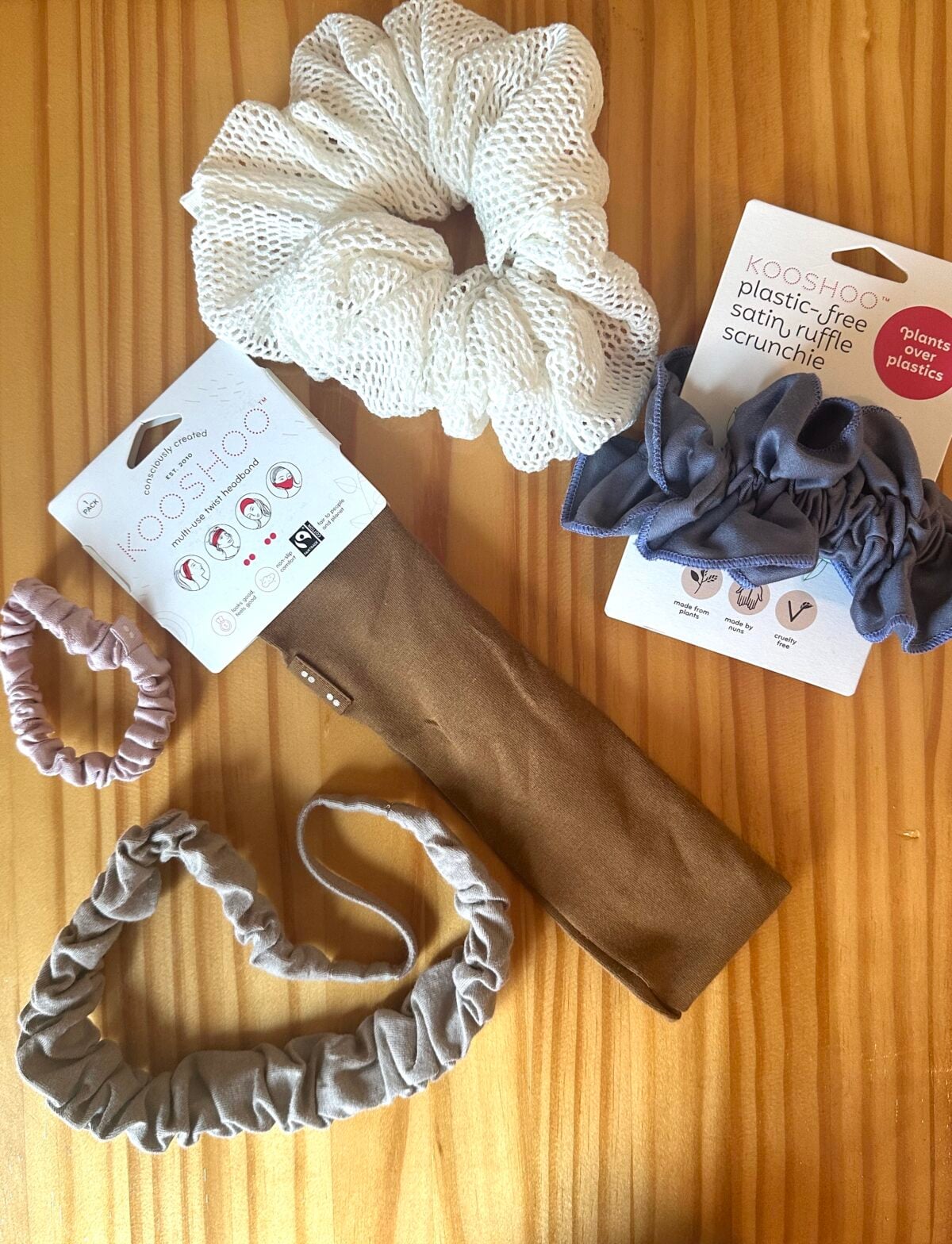 Assorted Kooshoo plastic-free hair accessories, including scrunchies and a brown headband, displayed on a wooden surface.