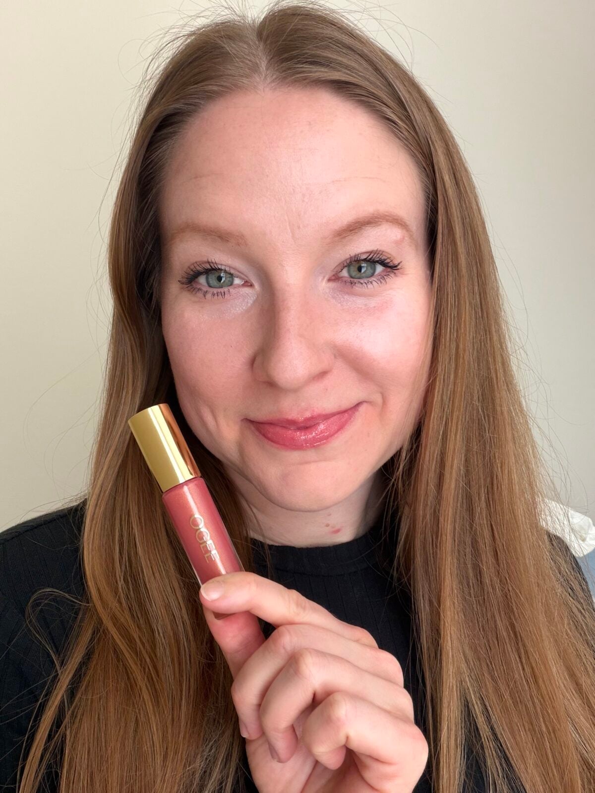 A smiling woman with long brown hair holds a tube of pink lip color in front of her face.
