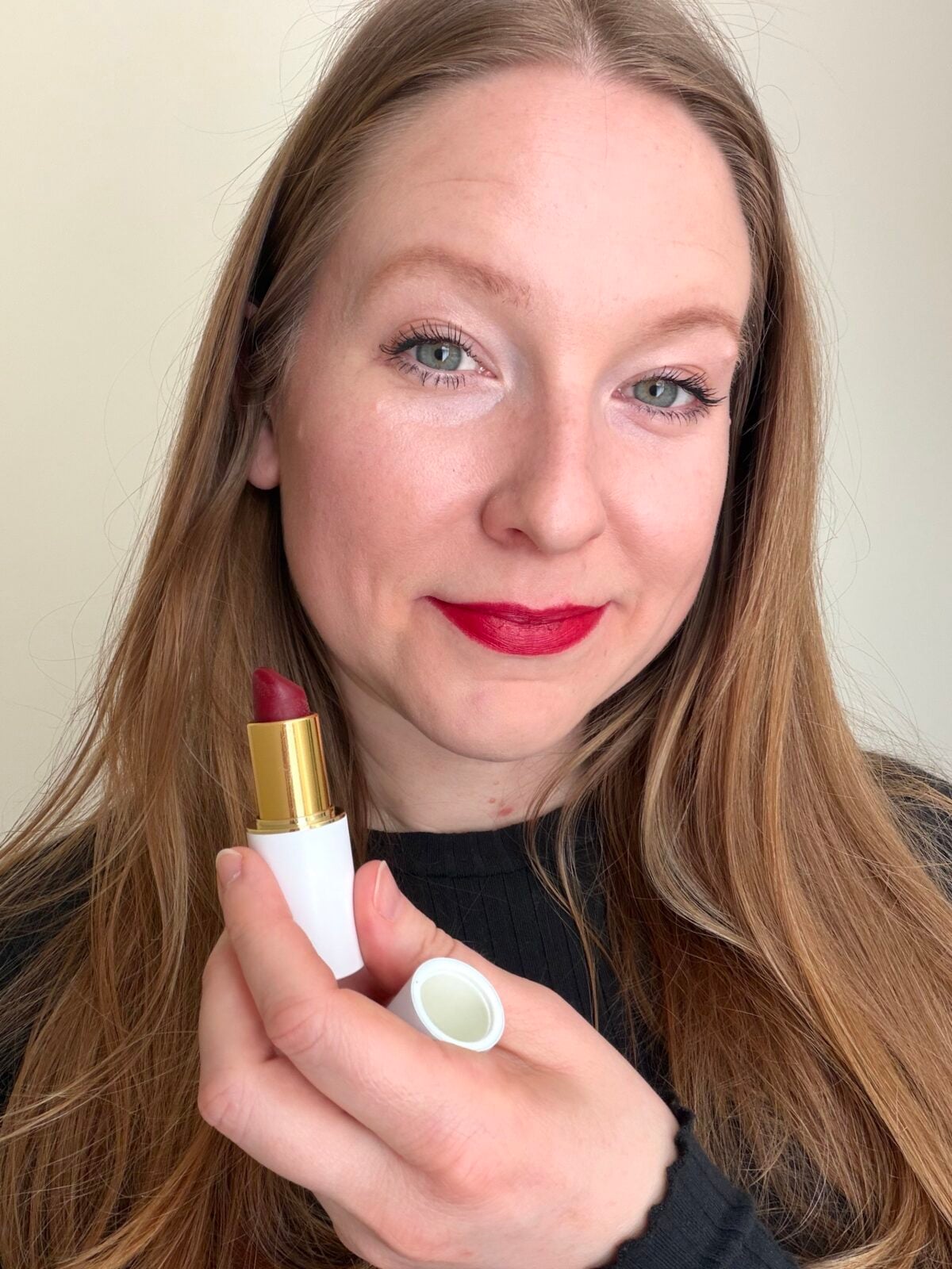 The photo shows a smiling woman with long light-brown hair and red lipstick holding an open tube of Ogee brand lipstick.