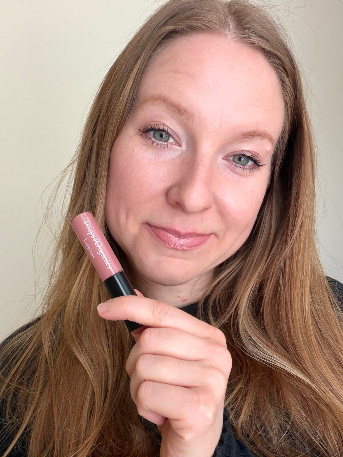 The photo shows a smiling woman with long brown hair holding a tube of pink lip color from the brand Toups & Co Organics in front of her face.