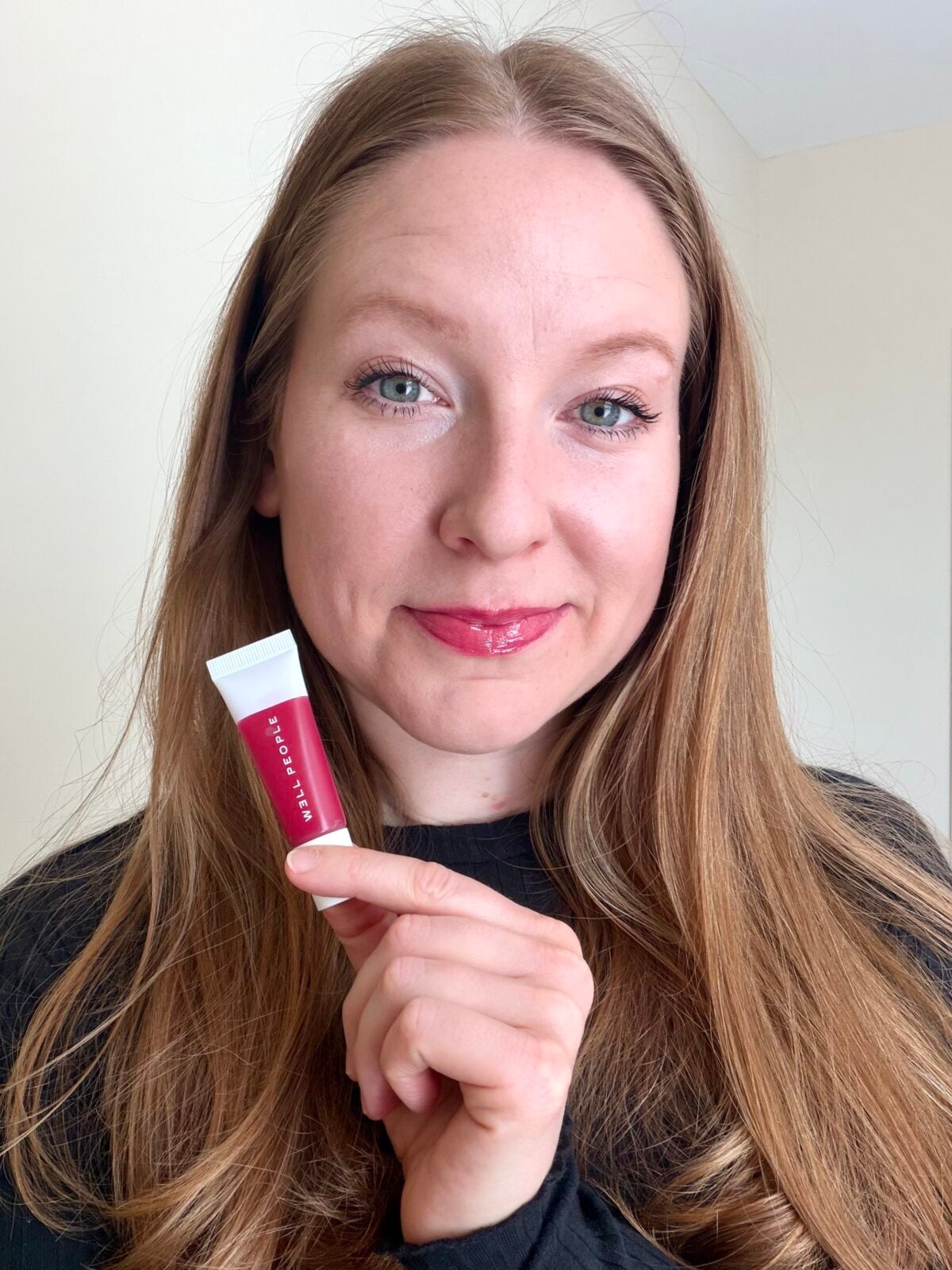 The photo shows a smiling woman with long light-brown hair holding a small tube of Well People Peptide Lip Balm in a berry shade.