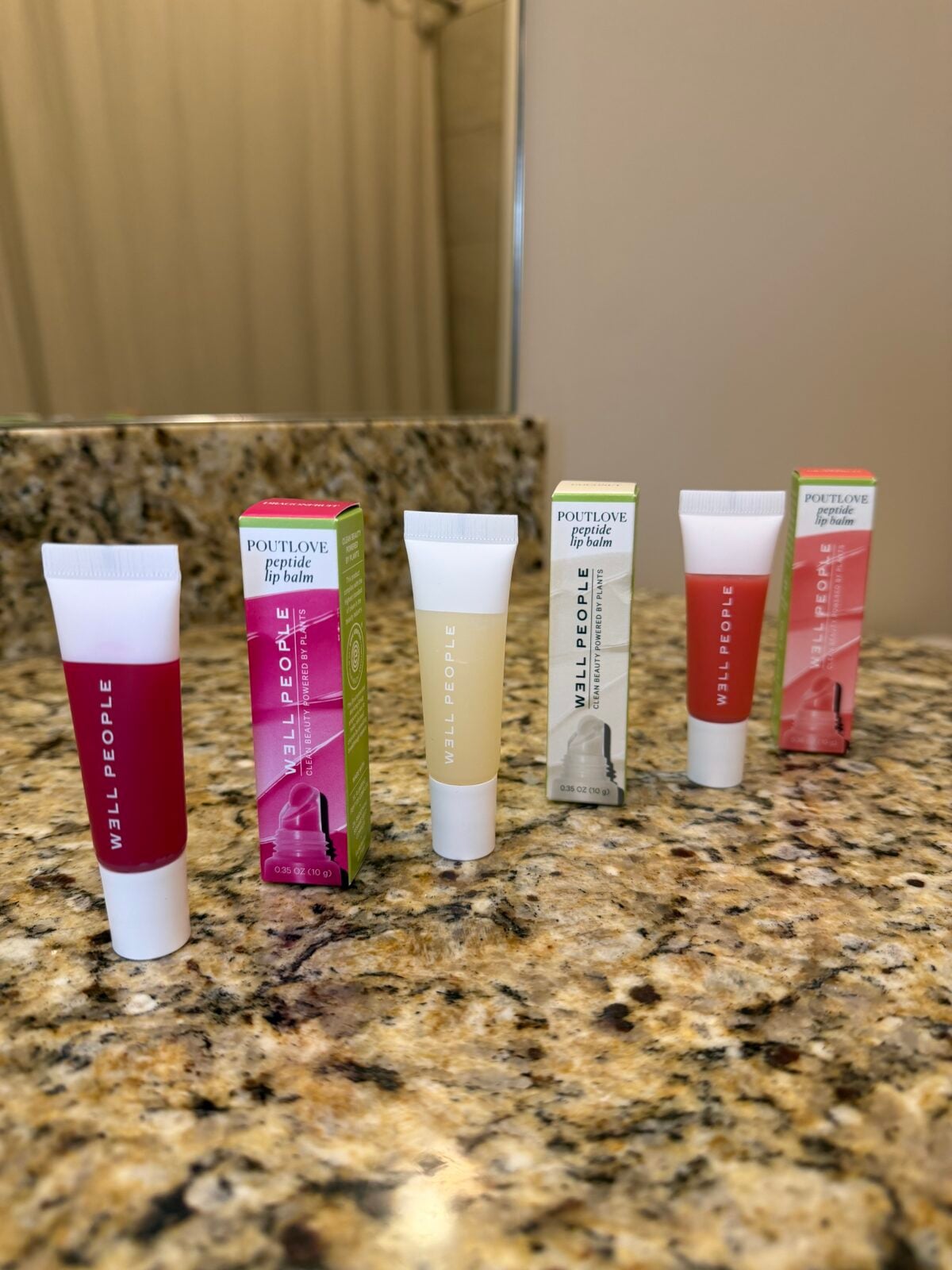 The photo displays three tubes and two boxes of Well People "Poutlove" Peptide Lip Balm in different shades arranged on a speckled granite countertop.