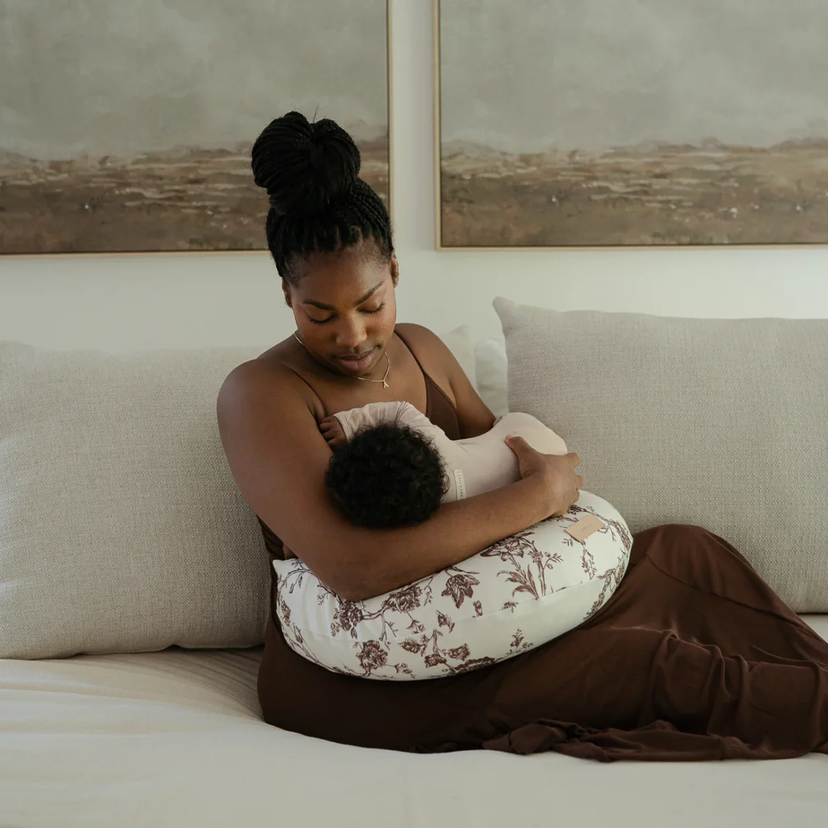 A woman in a brown dress sits on a bed and cradles her baby while using a floral-patterned nursing pillow for support in a room decorated with landscape paintings.