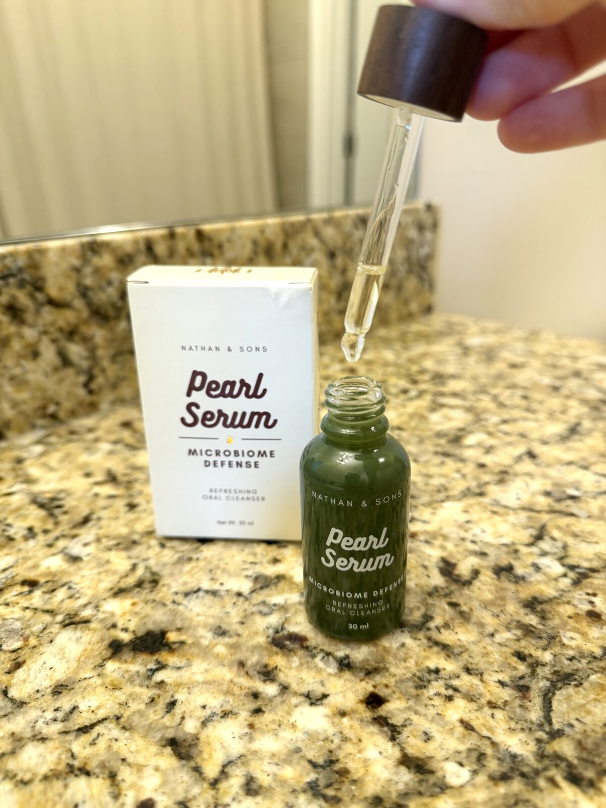 A hand holds a dropper above an open bottle of Nathan & Sons Pearl Serum on a granite bathroom counter, with the product box and reflection in the background.