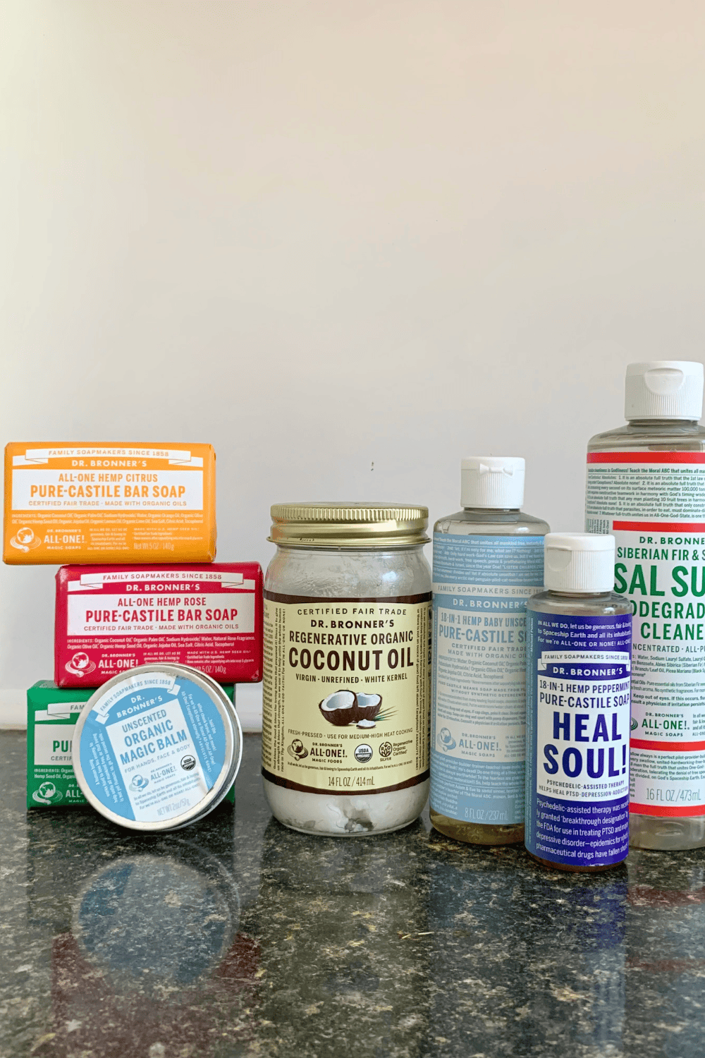 A collection of various Dr. Bronner's all-in-one products—including bar soaps, liquid soaps, organic balm, and coconut oil—are displayed on a dark countertop.