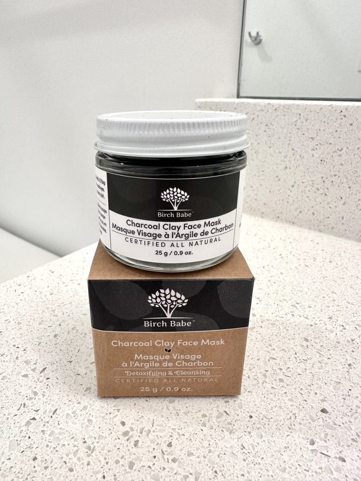 The photo shows a glass jar of Birch Babe "Charcoal Clay Face Mask" sitting on its brown cardboard packaging box on a white speckled bathroom countertop.