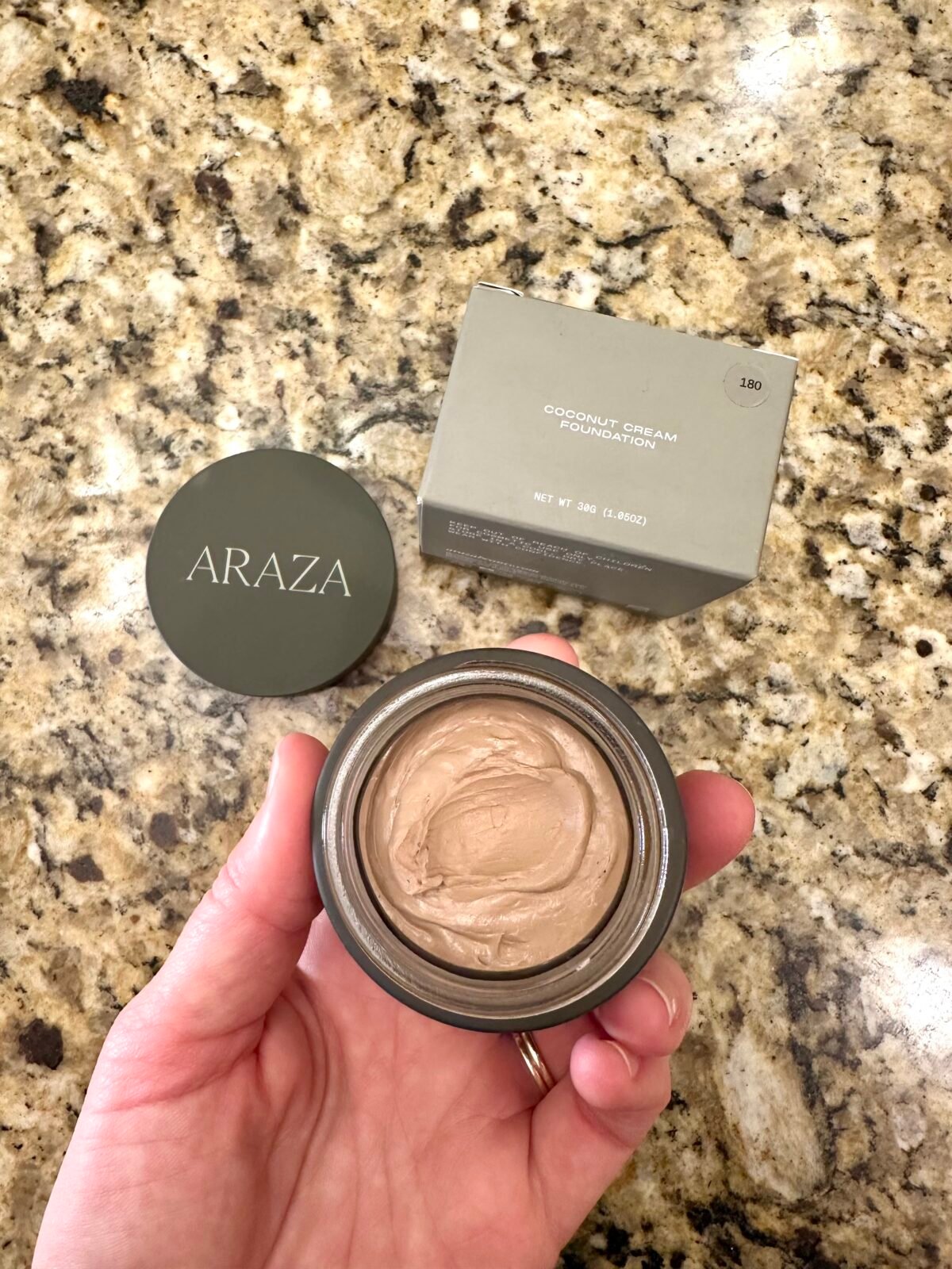 Hand holding an open jar of Araza Coconut Cream Foundation with the box and lid beside it on a granite countertop.