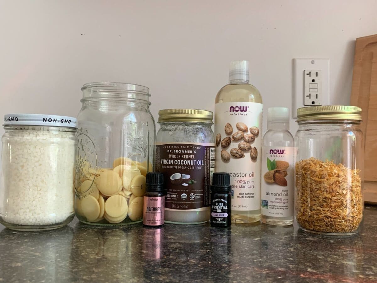 A countertop is shown with an array of jars and bottles containing ingredients such as castor oil, almond oil, coconut oil, cocoa butter wafers, and what appears to be calendula.