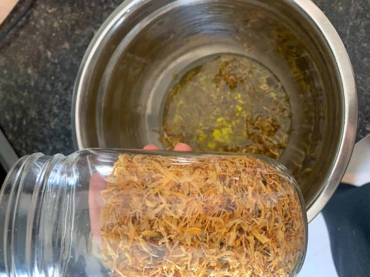 A jar filled with dried calendula is held over a metal bowl that contains a mixture of oil 