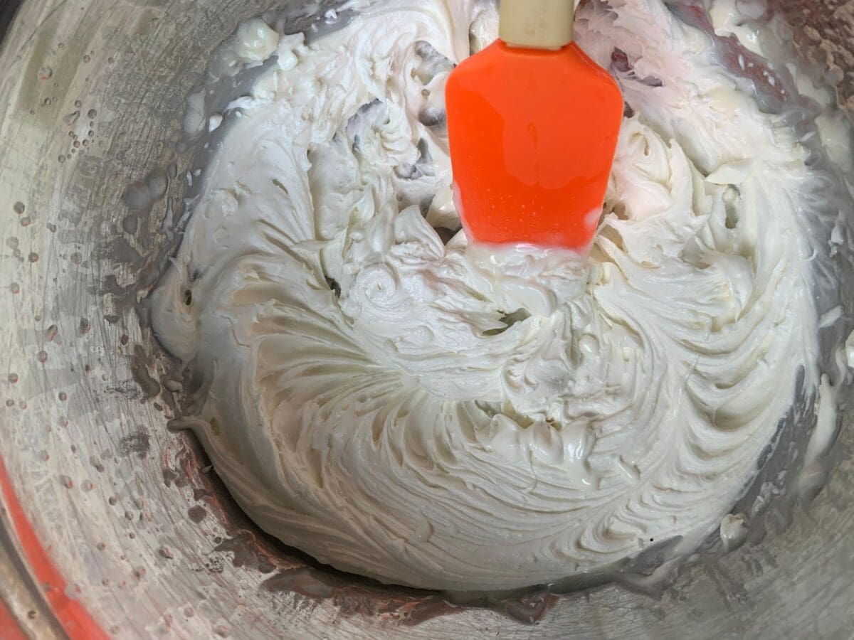 A spatula with an orange handle is shown in a metal bowl, mixing a white, creamy homemade lotion