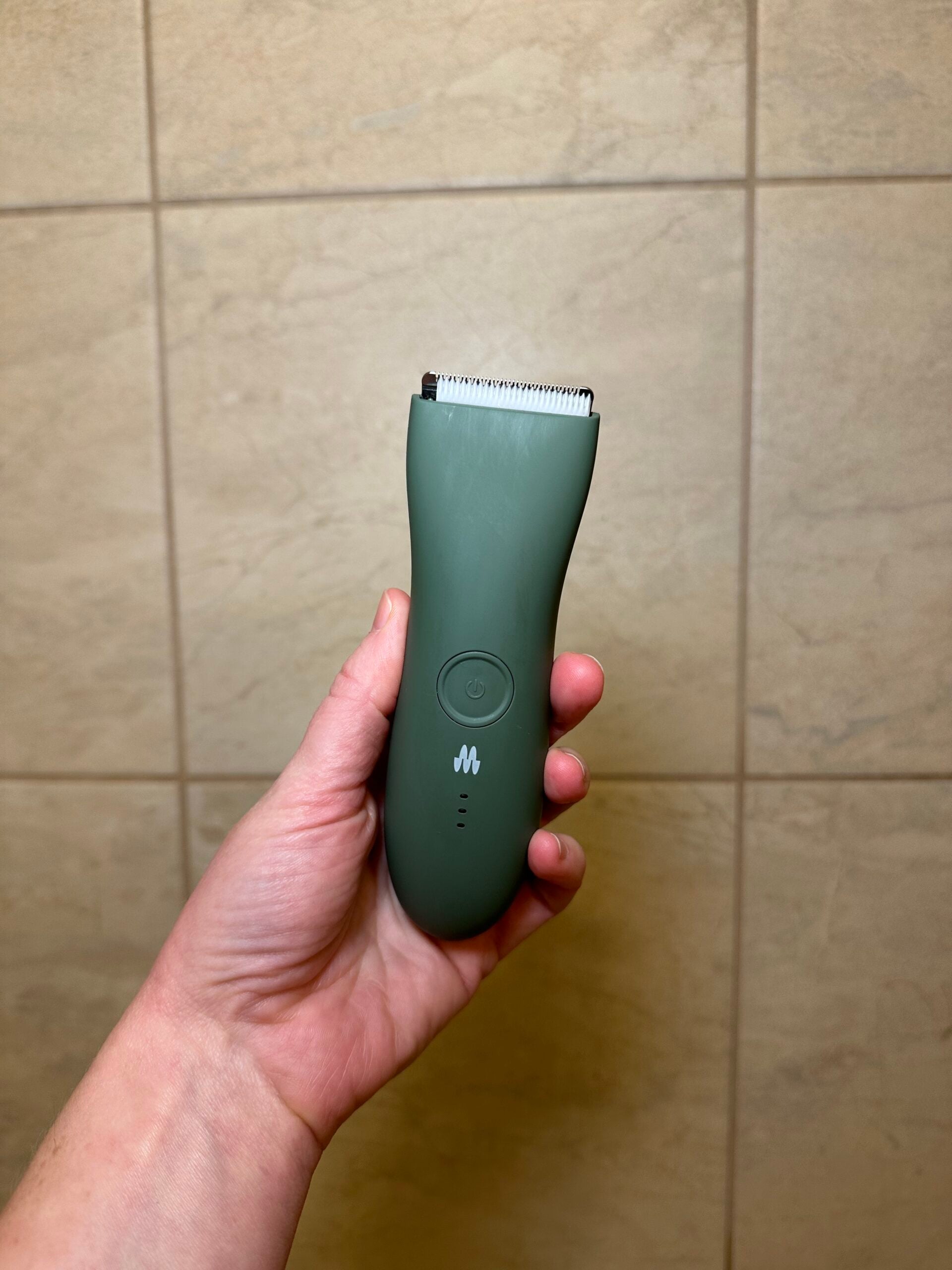 Hand holding a green Meridian body hair trimmer in front of beige tiled wall.