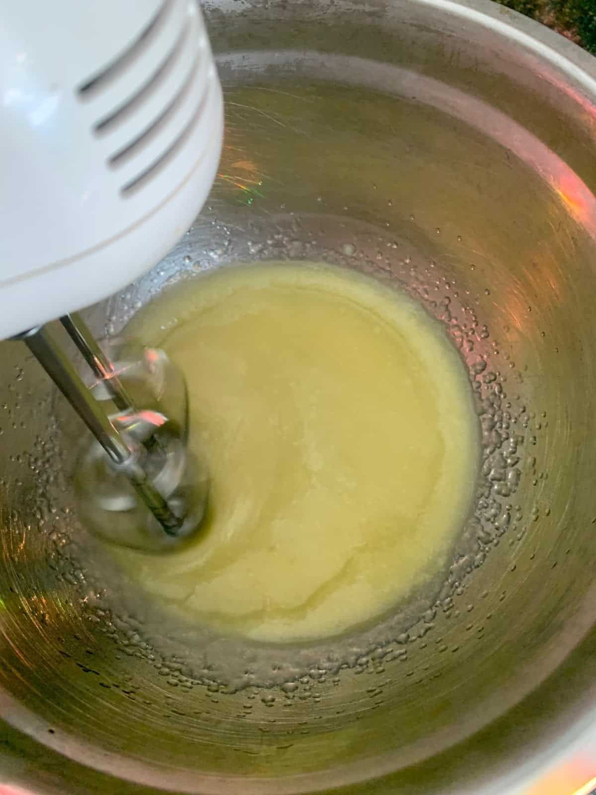 A white electric mixer is shown mixing a cacao butter, coconut and sweet almond oil in a silver metal bowl.
