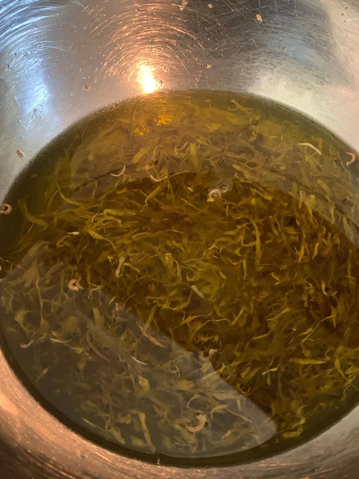 A close-up, top-down view shows a metal bowl filled with oil and herb infusion 