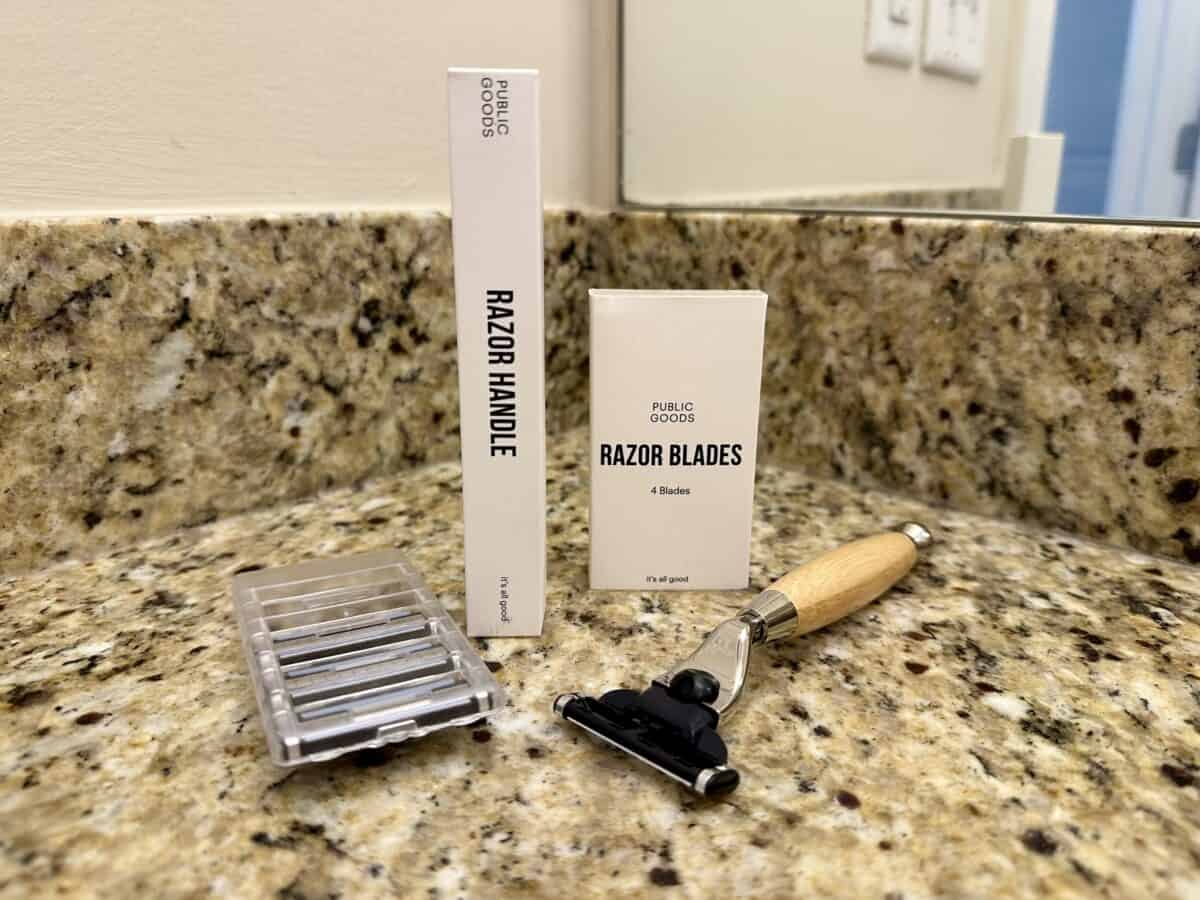 A Public Goods razor set displayed on a granite bathroom countertop, including a wooden-handled razor, a box labeled &ldquo;Razor Handle,&rdquo; a box labeled &ldquo;Razor Blades,&rdquo; and a plastic case holding replacement blades. A mirror and wall outlet are visible in the background.