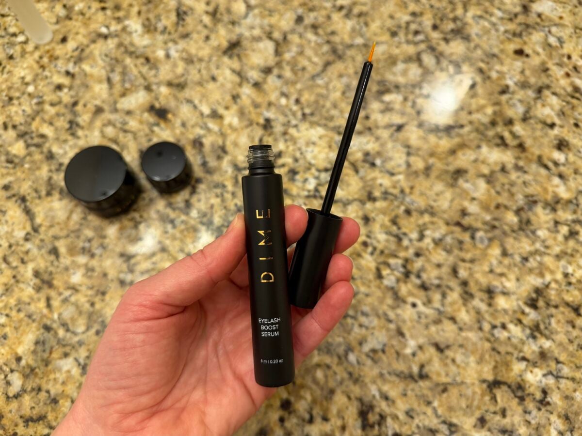 A hand holds an open tube of DIME Eyelash Boost Serum with the applicator wand exposed. Two black caps rest on a granite countertop in the background.