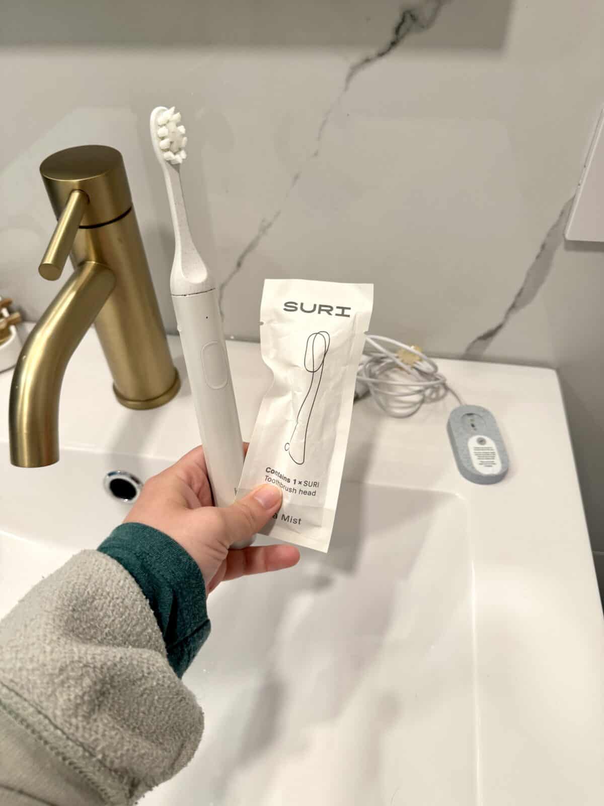 Hand holding a SURI electric toothbrush and a replacement brush head packet over a bathroom sink.