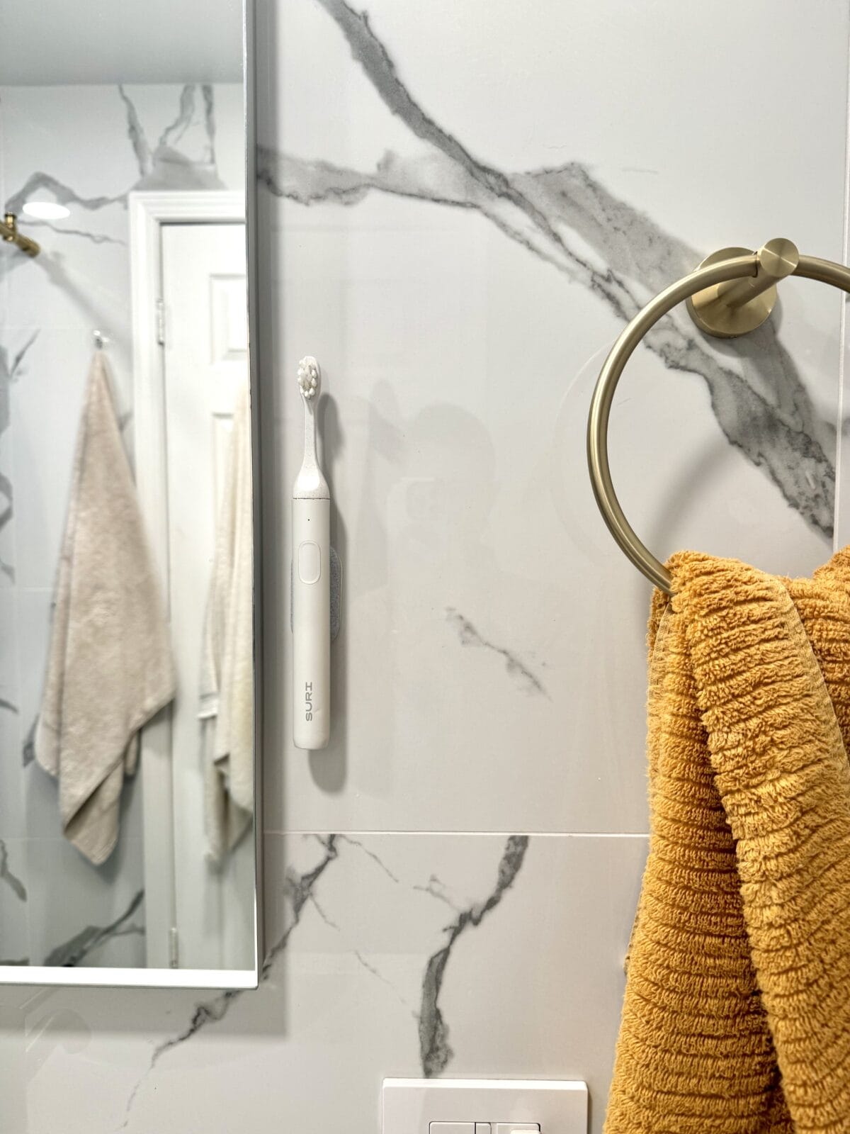 SURI electric toothbrush mounted on a bathroom wall next to a mustard yellow hand towel.