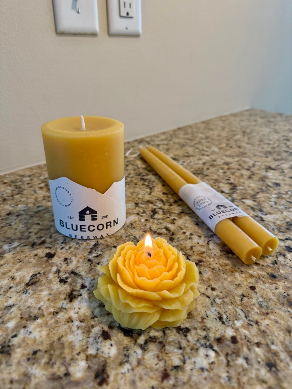 A yellow flower-shaped candle is lit on a granite countertop. Nearby are a yellow pillar candle labeled Bluecorn and a pair of yellow taper candles, also labeled Bluecorn. Electrical outlets are on the wall.