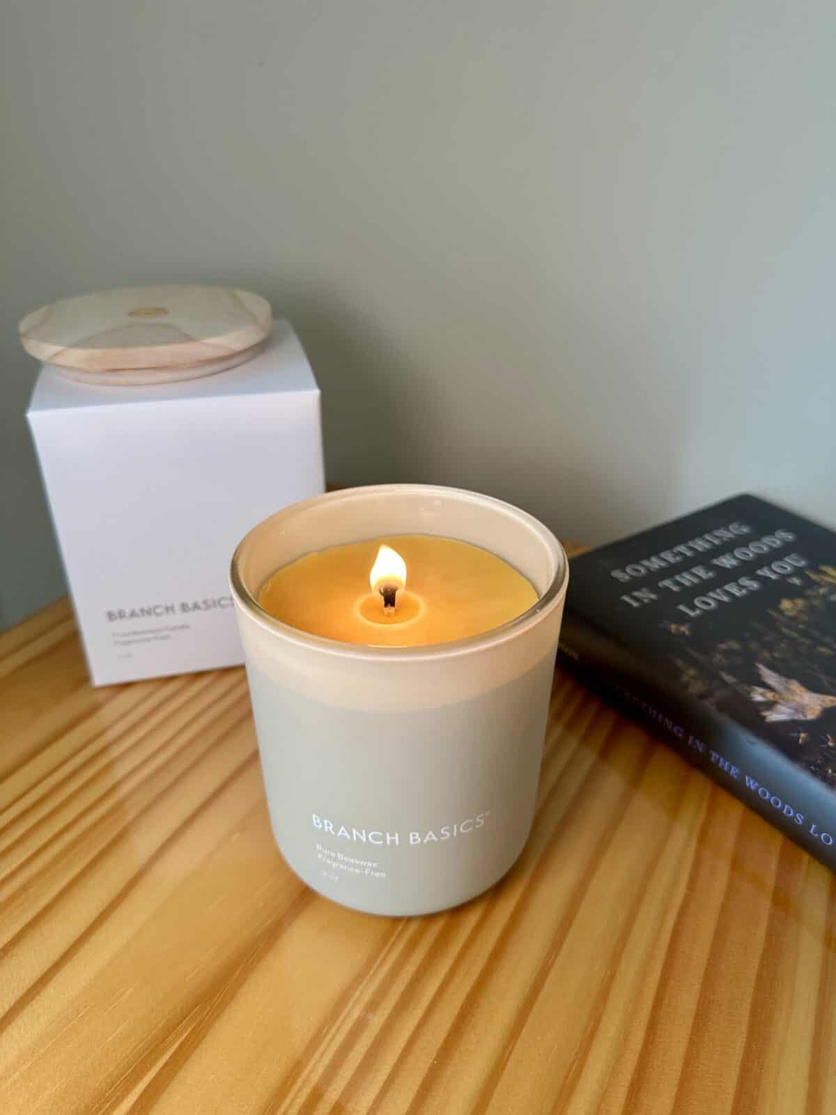 A lit Branch Basics pure beeswax candle in a matte glass jar sits on a wooden surface next to its box and a book titled Something in the Woods is Loves You.