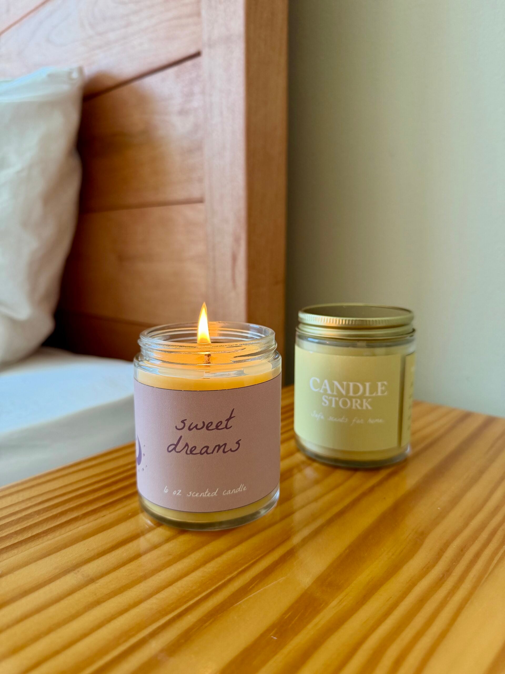 A lit candle labeled sweet dreams sits on a wooden nightstand next to an unopened candle jar labeled CANDLE STORK. A bed with a white pillow and wooden headboard is partially visible in the background.