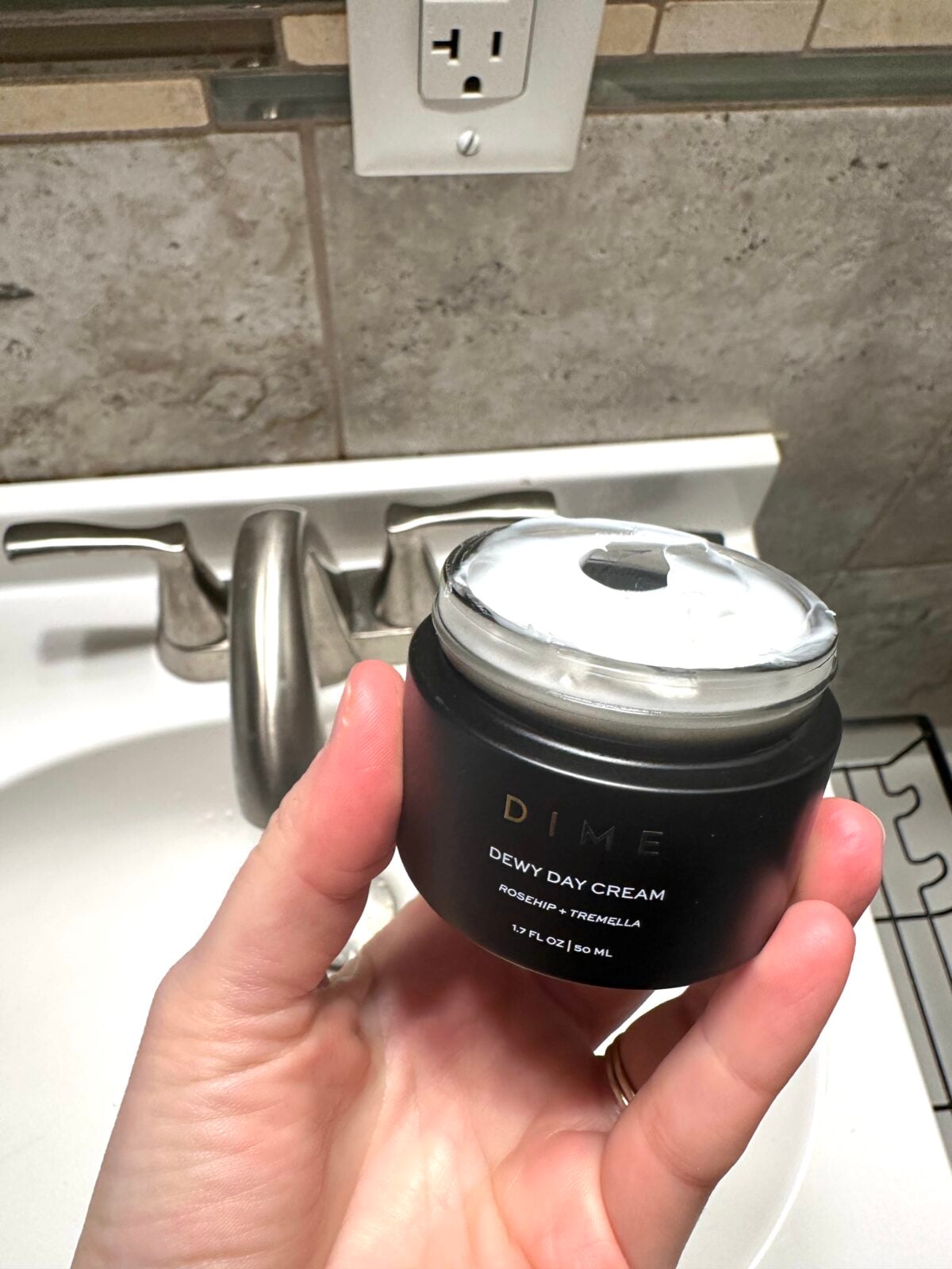 A hand is holding an open jar of DIME Dewy Day Cream above a white bathroom sink with a silver faucet. A power outlet and tiled wall are visible in the background.