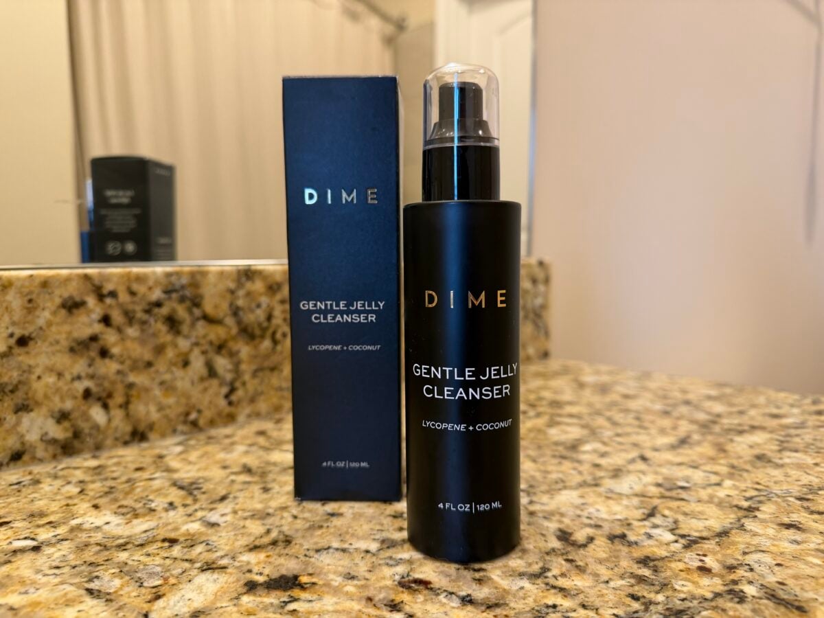 A black bottle of DIME Gentle Jelly Cleanser with lycopene and coconut stands on a granite countertop next to its matching box. A bathroom mirror and other products are visible in the background.