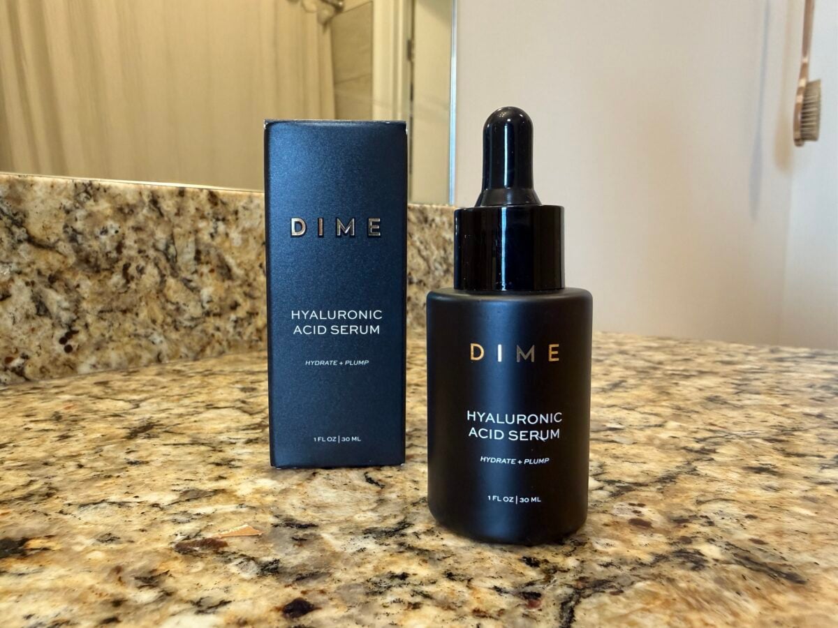 A black bottle of DIME Hyaluronic Acid Serum with a dropper sits on a granite bathroom counter next to its matching black box packaging. A sink and a portion of a mirror are visible in the background.