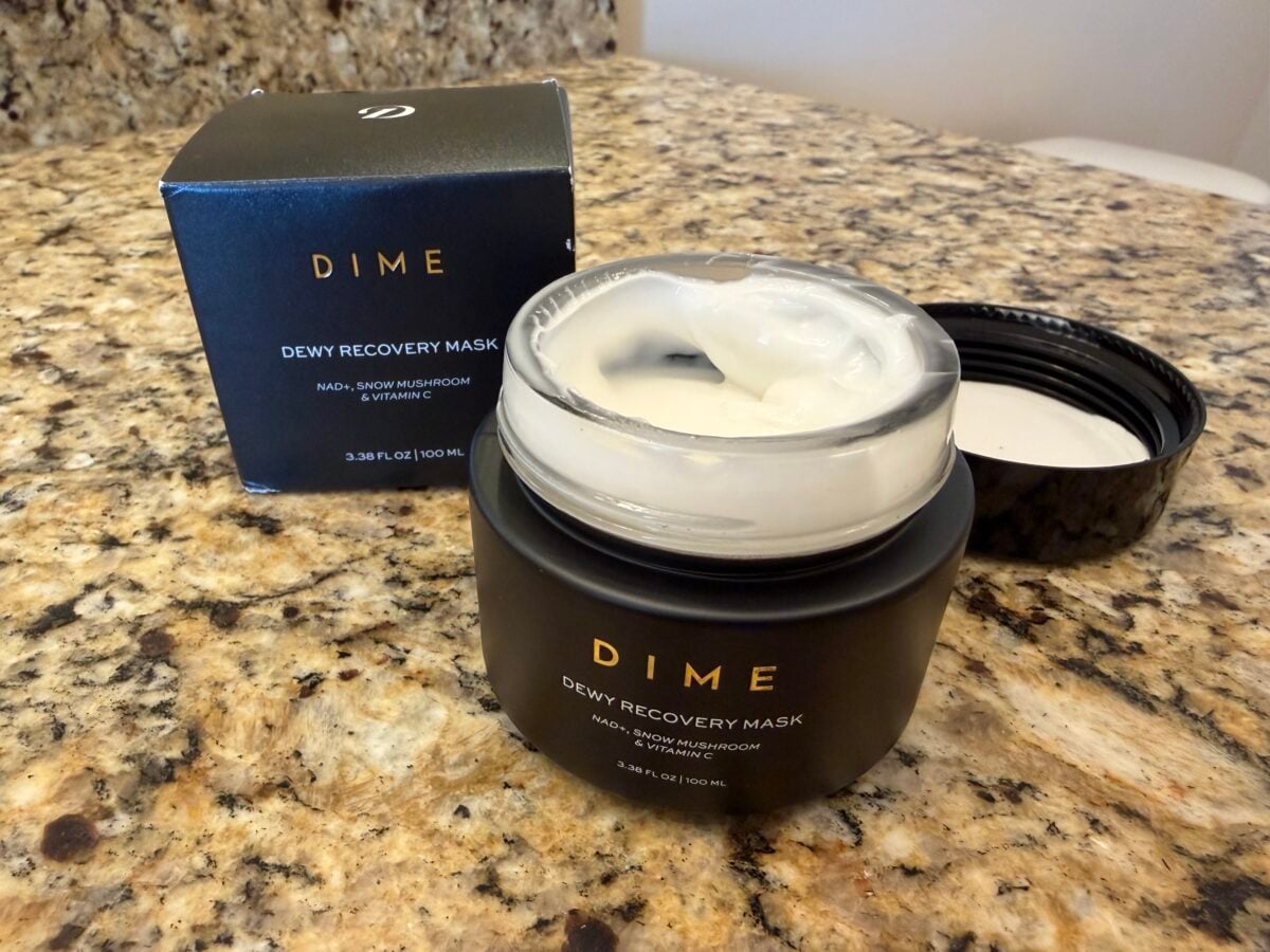 An open jar of Dime Dewy Recovery Mask with its lid placed nearby, next to its black product box, all sitting on a speckled granite countertop. The jar is partially filled with thick white cream.